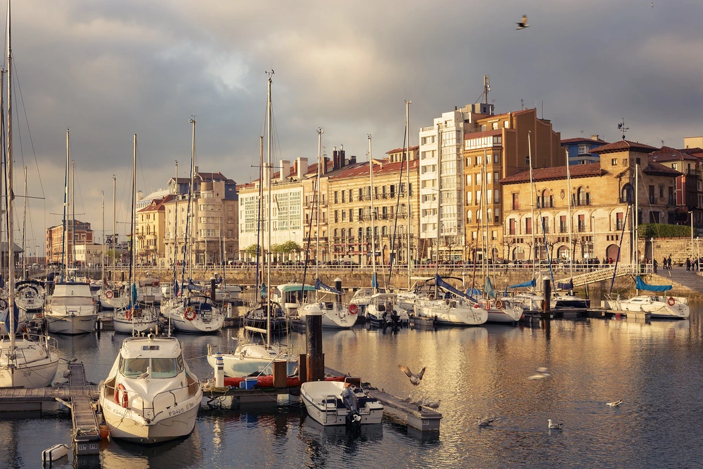 Barcos amarrados en el Puerto Deportivo de Gijón con los edificios de la ciudad al fondo, un lugar perfecto para terminar tu ruta por el norte de España.