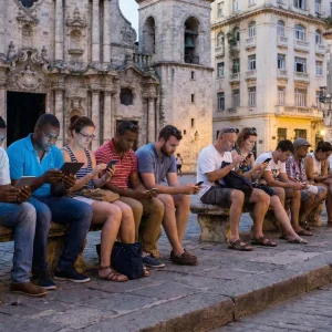 Fila de personas, incluyendo turistas y locales, sentadas en bancos de piedra en una plaza colonial al atardecer, con sus rostros iluminados por las pantallas de sus móviles mientras usan un punto de conexión Wifi público